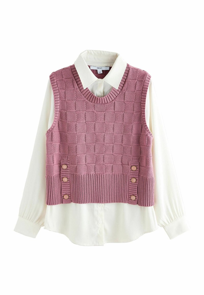 Chemise crème à manches longues avec col sous un gilet sans manches en maille rose à motif à carreaux et boutons décoratifs près de l'ourlet.