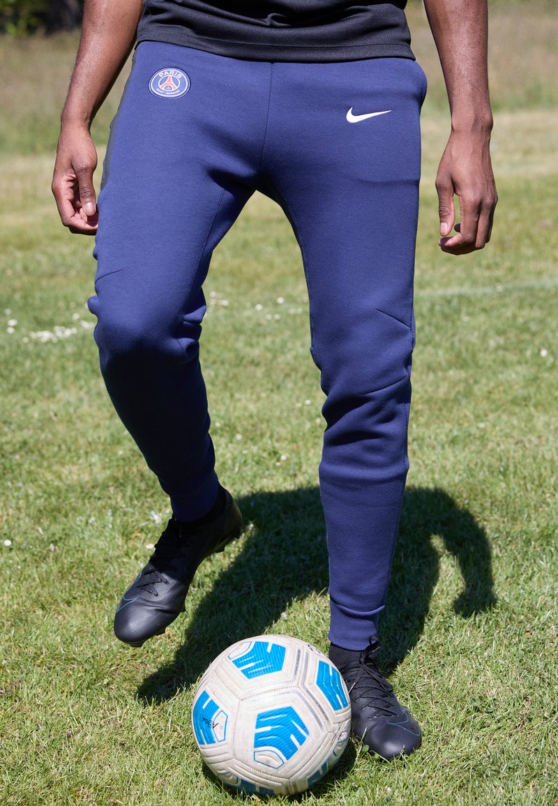 Jogging athlétiques marins avec un design ajusté et des poignets élastiques, arborant un logo du Paris Saint-Germain et un swoosh Nike, associés à des chaussures noires.