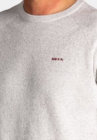 Close-up van een lichtgrijze textuur gebreid trui met een ronde halslijn en kleine rode geborduurde letters "N.Z.A." op de borst.