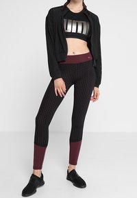 Atletyczna kobieta ubrana w czarny krótki top sportowy, czarną perforowaną kurtkę, wzorzyste legginsy w kolorze czarnym i bordowym oraz czarne sneakersy, stojąca.