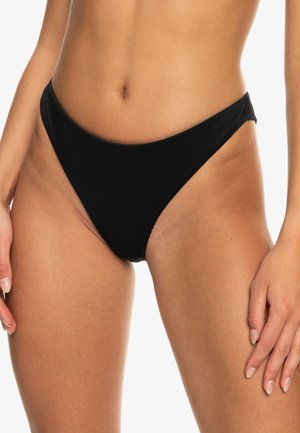 Närbild av en kvinna som bär högskurna svarta ribbade bikinibyxor med händerna vilande vid höfterna mot en vit bakgrund.