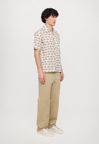 Kurzarmhemd aus cremefarbenem Stoff mit einem bunten Druck von verschiedenen Objekten. Kombiniert mit beigen, weiten Hosen und hellen Sneakers.