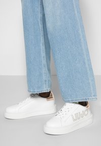 Valkoiset nahkakengät, joissa on texturoitu vaaleanbeige yksityiskohta kantapäässä ja "LIU JO" -logo, yhdistettynä vaaleansiniset farkkulegginsit jotka ovat leveät lahkeet.