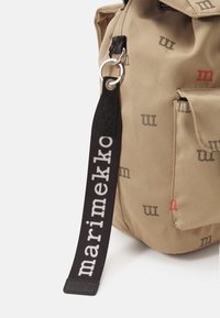 Borsa in tessuto beige con motivi logo sottili in grigio e rosso. Manico nero con "marimekko" in lettere bianche; dettaglio con anello metallico rinforzato.