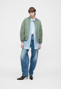 Asmuo stovi dėvėdamas žalią bomberį, mėlyną dryžuotą marškinėlį, plačias kelnes ir juodas smailiąsias kulniukus prieš vientisą foną.