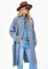 Kobieta w brązowym kapeluszu, dużych okularach przeciwsłonecznych, szarym długim płaszczu na niebieskiej dżinsowej kurtce i jasnoniebieskich dżinsach, stojąca z jedną ręką w kieszeni.