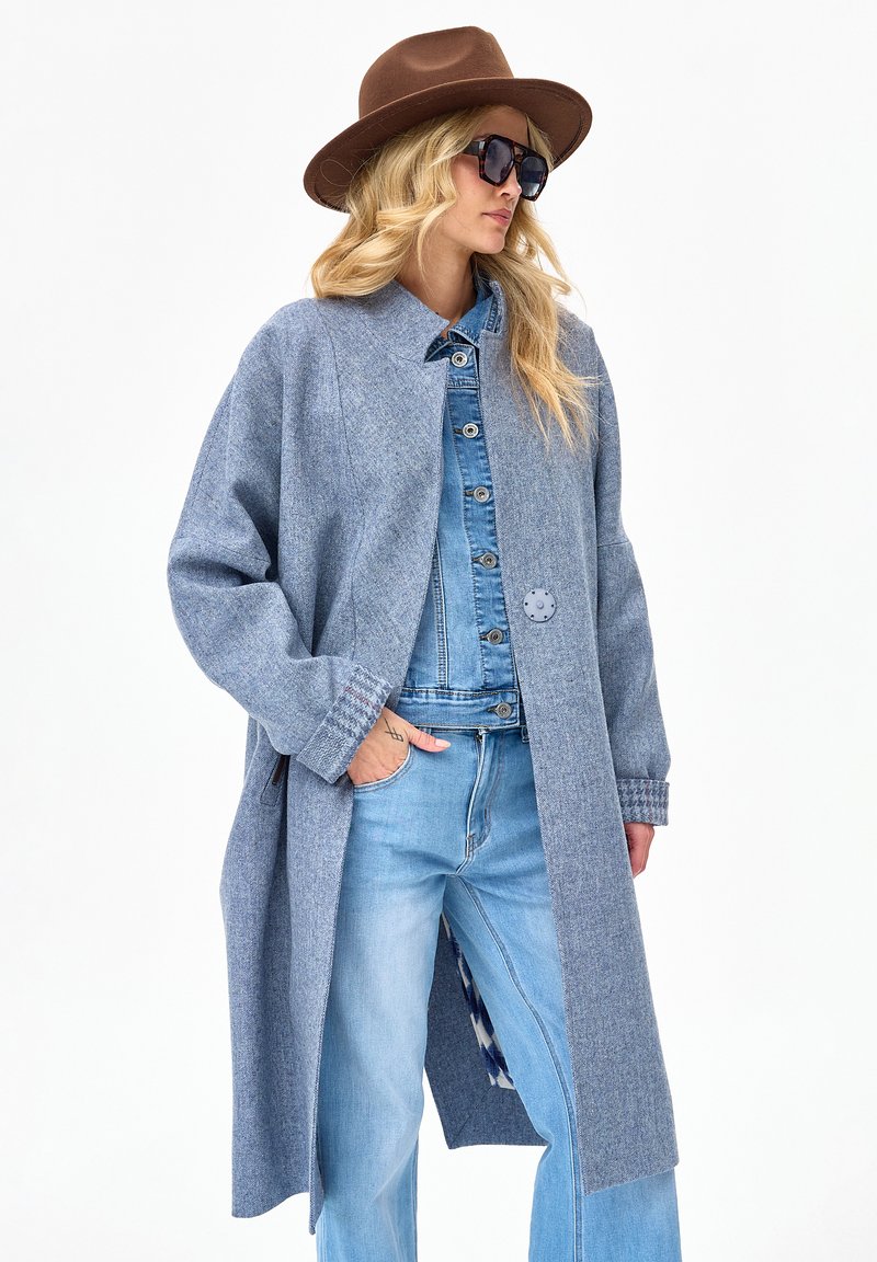 Kobieta w brązowym kapeluszu, dużych okularach przeciwsłonecznych, szarym długim płaszczu na niebieskiej dżinsowej kurtce i jasnoniebieskich dżinsach, stojąca z jedną ręką w kieszeni.