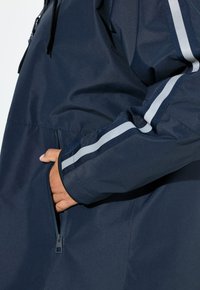 Chaqueta cortavientos navy con un bolsillo con cremallera, franjas blancas en contraste en las mangas y un tejido ligero y texturizado.
