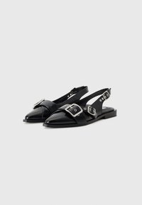 Black leather slingback shoes with pointed toes, crisscross straps, large silver buckles, and flat soles. Smooth texture and minimal design.