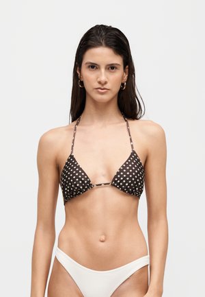 Mladá žena s dlhými tmavými vlasmi nosí hnedý vzorovaný bikiny top v tvare trojuholníka a biele bikiny nohavičky na jednoduchom pozadí.