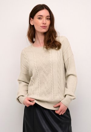 Beige Kabelstrickpullover mit strukturiertem Design, rundem Halsausschnitt und gerippten Bündchen, kombiniert mit einem schwarzen Lederrock.