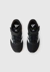 Chaussures de sport noires avec un dessus en mesh, dotées de bandes blanches et d'une bride Velcro. La semelle est blanche avec un motif texturé.