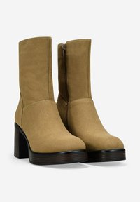 Par de botas de gamuza color marrón claro hasta la mitad de la pantorrilla con cremallera lateral, punta redonda y gruesos tacones de bloque de madera apilada sobre fondo blanco.