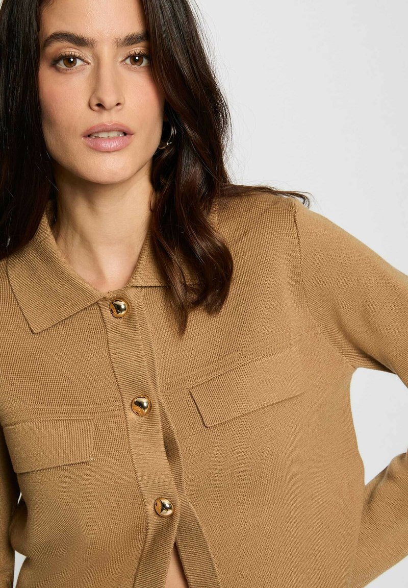 Cardigan marrone lavorato a maglia con design colletto, bottoni dorati e due tasche frontali. La texture è a coste, offrendo un aspetto accogliente.