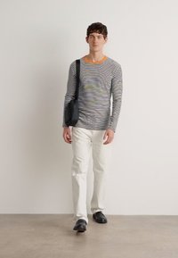 Camicia a righe a maniche lunghe con scollo arancione, pantaloni bianchi e borsa nera. Modella in piedi contro uno sfondo neutro. Dettagli del tessuto texturizzati visibili.