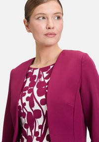 Fuksjowy żakiet z wydłużonym klapami, w połączeniu z białą bluzką z intensywnym bordowym kwiatowym wzorem. Gładki materiał i dopasowany krój.