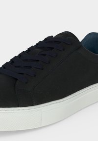 Sort lavtop sneaker med mørke snørebånd og hvid gummisål, der viser side- og topdetaljer på en ensartet lys baggrund.