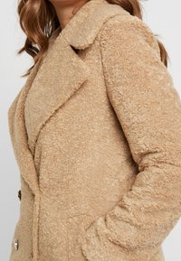 Cappotto teddy beige con colletto a punte, tessuto testurizzato, design doppiopetto e bottoni dorati sulla parte anteriore.