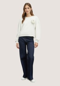 Suéter blanco con un acabado texturizado y una gran flor de tela en el lado izquierdo del pecho, combinado con jeans de pierna ancha azul oscuro y zapatillas blancas.