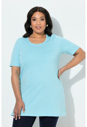 Kobieta o ciemnych włosach, nosząca złote kolczyki w kształcie koła, jasnoniebieską tunikę z krótkim rękawem i ciemne spodnie, stojąca z jedną ręką na biodrze.