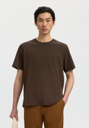 Giovane uomo che indossa una t-shirt marrone a maniche corte e pantaloni marroni, in piedi con una mano in tasca e l'altra che tiene la tracolla di una borsa beige.