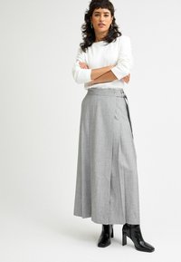 Grauer, langer, weiter Rock aus strukturiertem Stoff mit asymmetrischem Design und seitlichen Knöpfen, kombiniert mit schwarzen, eckigen Heels.