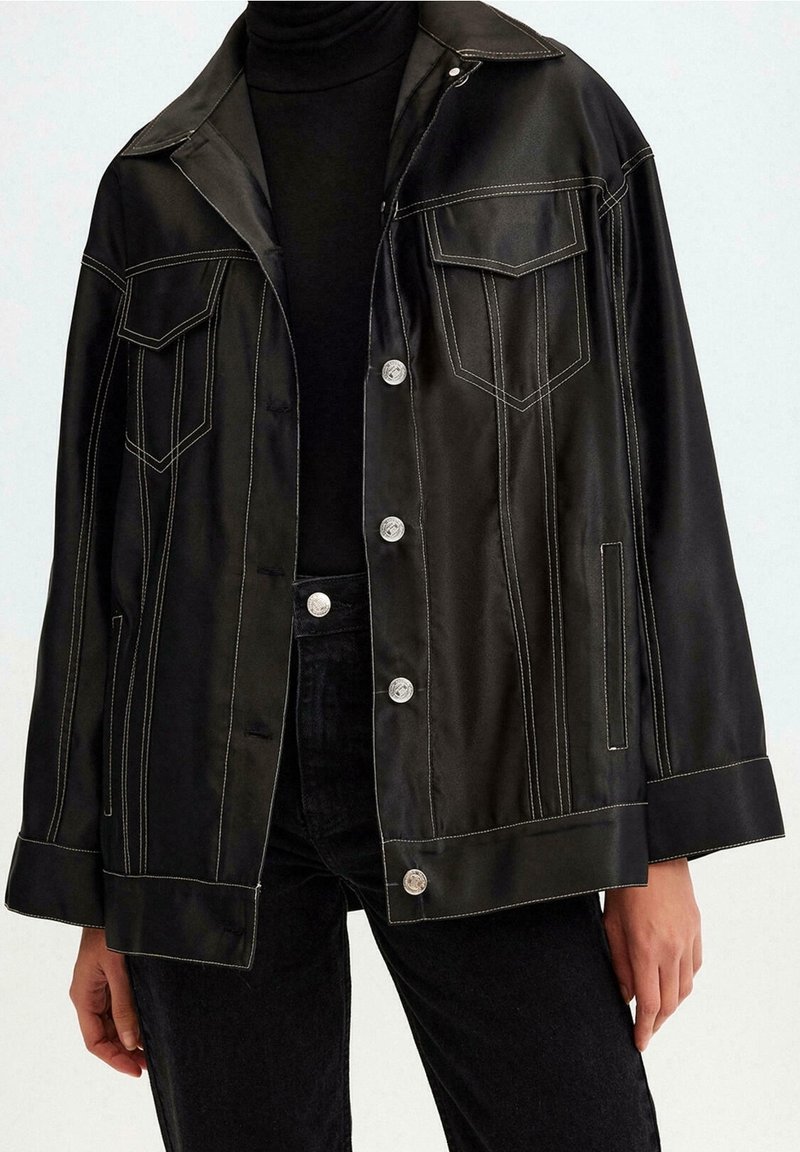 Zwarte satijnen jack met een grote kraag, twee borstzakken en contrasterende witte stiksels. Voorzien van zilveren knopen en een relaxte pasvorm.