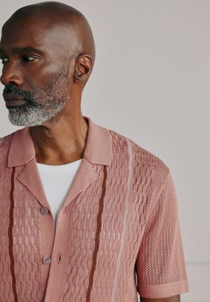 Vīrietis ar pelēku bārdu, valkājot rozā rakstainu īsroku krekla kreklu pār baltu ribotu krekliņu, skatās pa kreisi.