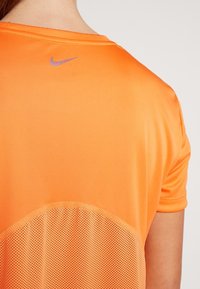 Oranžové sportovní tričko z hladké, lehké látky s síťovaným panelem ve spodní části. Malé fialové logo Nike se nachází u výstřihu.