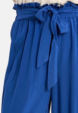 Pantaloni blu a vita alta con vita arricciata e cintura in tessuto annodata a fiocco sul davanti, indossati con una maglietta beige chiaro.