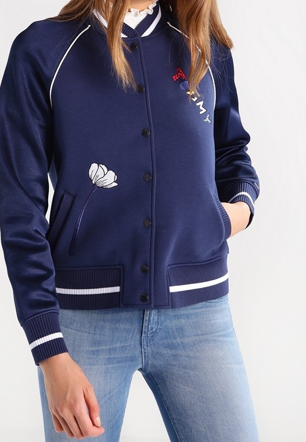 Femme portant une veste bombers bleu marine boutonnée avec une broderie florale blanche, des poignets rayés et un jean bleu clair.