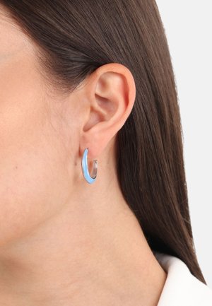 Pendiente de aro de plata con esmalte azul, que presenta un diseño curvado. Textura suave con un acabado brillante, posicionado sobre un tono de piel claro.