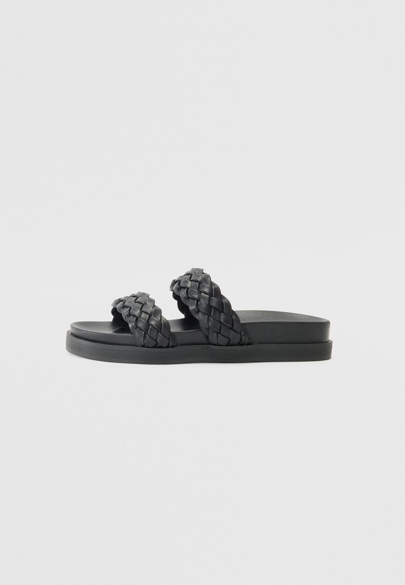 Svarte sandaler har to flettede stropper og en flat, teksturert såle. Designet er minimalistisk med en glatt overflate og uten dekorasjoner.