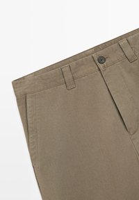 Beżowe spodnie wykonane z fakturowanej tkaniny, posiadające płaski przód, szlufki na pasek oraz pojedynczy czarny guzik w pasie.