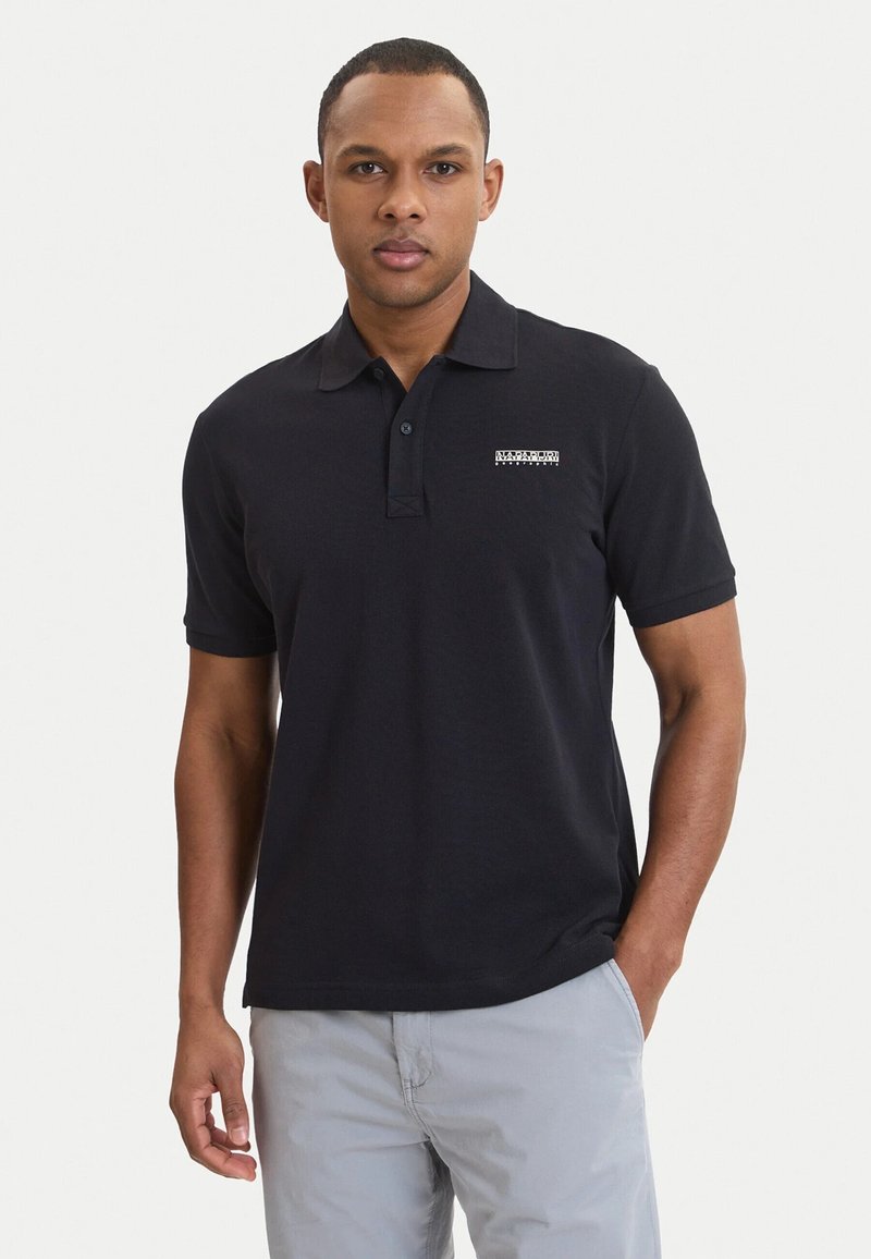 Mann trägt ein schwarzes kurzärmeliges Poloshirt mit kleinem Logo auf der Brust und hellgraue Hose, steht vor weißem Hintergrund.