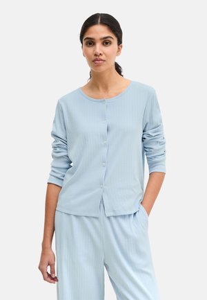 Frau mit zurückgebundenem dunklem Haar, die eine hellblaue, gerippte, knöpfbare Strickjacke und passende Hose trägt, mit einer Hand in der Tasche.