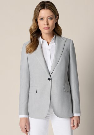 Kobieta o falowanych, jasnobrązowych włosach, ubrana w jasnoszary żakiet na białą, zapinaną na guziki koszulę i białe spodnie, stojąca na beżowym tle.