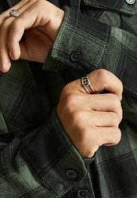 Camicia di flanella a scacchi neri e verdi con tessuto testurizzato, polsini con bottoni e anelli d'argento visibili sulle mani.