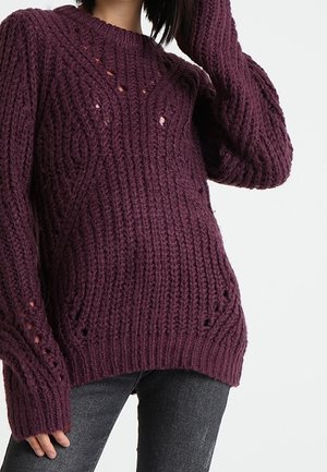 Kobieta ubrana w luźny, grubo dziergany fioletowy sweter z teksturowanymi wzorami oraz ciemnoszare dżinsy, pozująca z uniesioną jedną ręką.