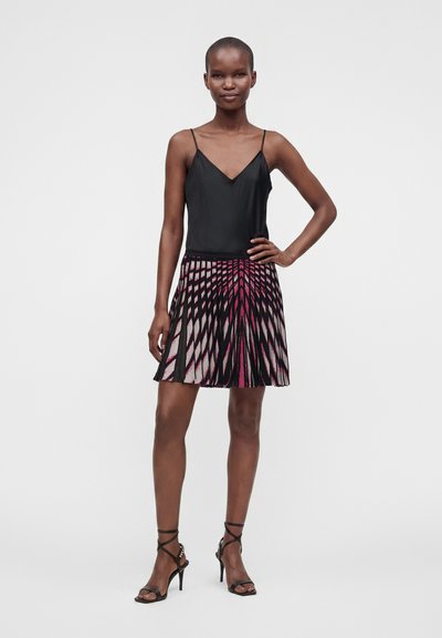 Femme debout, main sur la hanche, portant un débardeur noir, une jupe rose et noire à motifs et des sandales à talons noirs sur un fond uni.