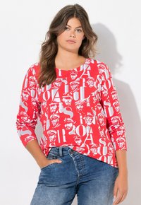 Rotes Langarmshirt mit grafischem Druck von Gesichtern und Buchstaben in Weiß. Aus Baumwollmaterial, mit rundem Ausschnitt, lockerer Passform, kombiniert mit blauen Jeans.