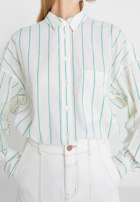 Camicia bianca con righe verticali turchesi, dotata di abbottonatura frontale, colletto e una tasca sul petto; realizzata in tessuto leggero.