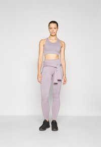 Soutien-gorge de sport lilas et legging taille haute assorti, avec un détail de taille nouée. Porté avec des chaussures de sport noires, texture lisse.