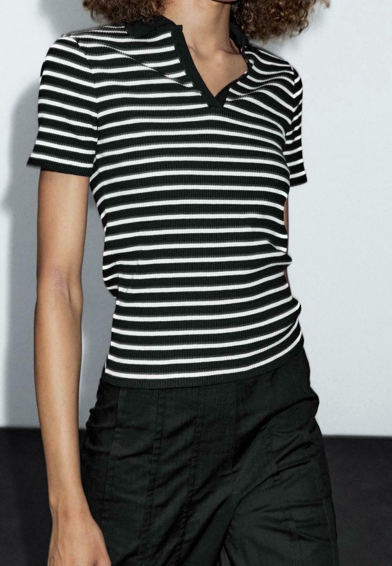 Personne portant une chemise à manches courtes à rayures horizontales noires et blanches et un pantalon noir taille haute sur un fond uni.