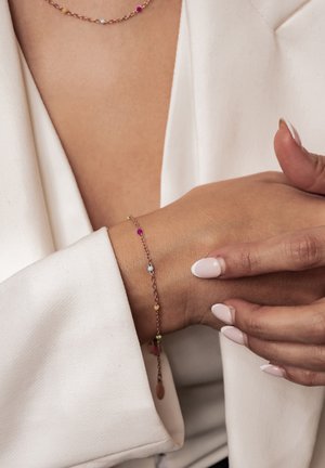 Zartes Goldkettchenarmband mit kleinen bunten Perlen, am Handgelenk getragen, kombiniert mit passender Halskette und weißem Blazerärmel.