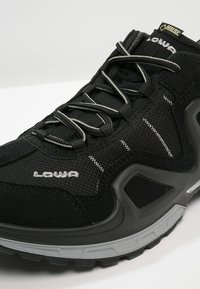 Schwarzer Sneaker mit einem Obermaterial aus Mesh und Wildleder, reflektierenden Akzenten, einer kontrastierenden grauen Gummisohle und einem Schnürsystem für einen sicheren Sitz.