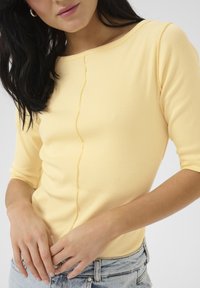 Top giallo a coste con scollatura a barca e maniche a tre quarti. Presenta una cucitura centrale e una texture morbida, abbinato a pantaloni corti in denim chiaro.
