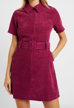 Kobieta w różowej, teksturowanej sukience ze sztruksu z krótkim rękawem i kołnierzem, z dopasowanym paskiem i złotą bransoletką na prawym nadgarstku.