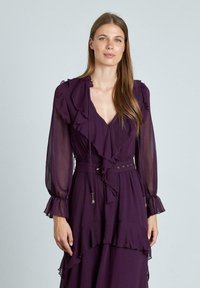 Vestido púrpura hecho de un tejido ligero que presenta acentos de volantes, mangas largas y transparentes, una cintura ceñida con cinturón y un escote en V profundo.