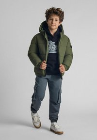 Giacca a vento verde con cappuccio, felpa blu navy con logo, pantaloni cargo grigi e sneakers con dettagli chiari. Look casual e a strati.